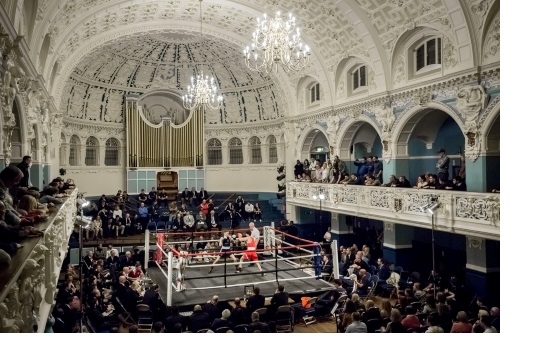 A view from the balcony of a hall, in the centre of which is a boxing ring, a fight in progress.