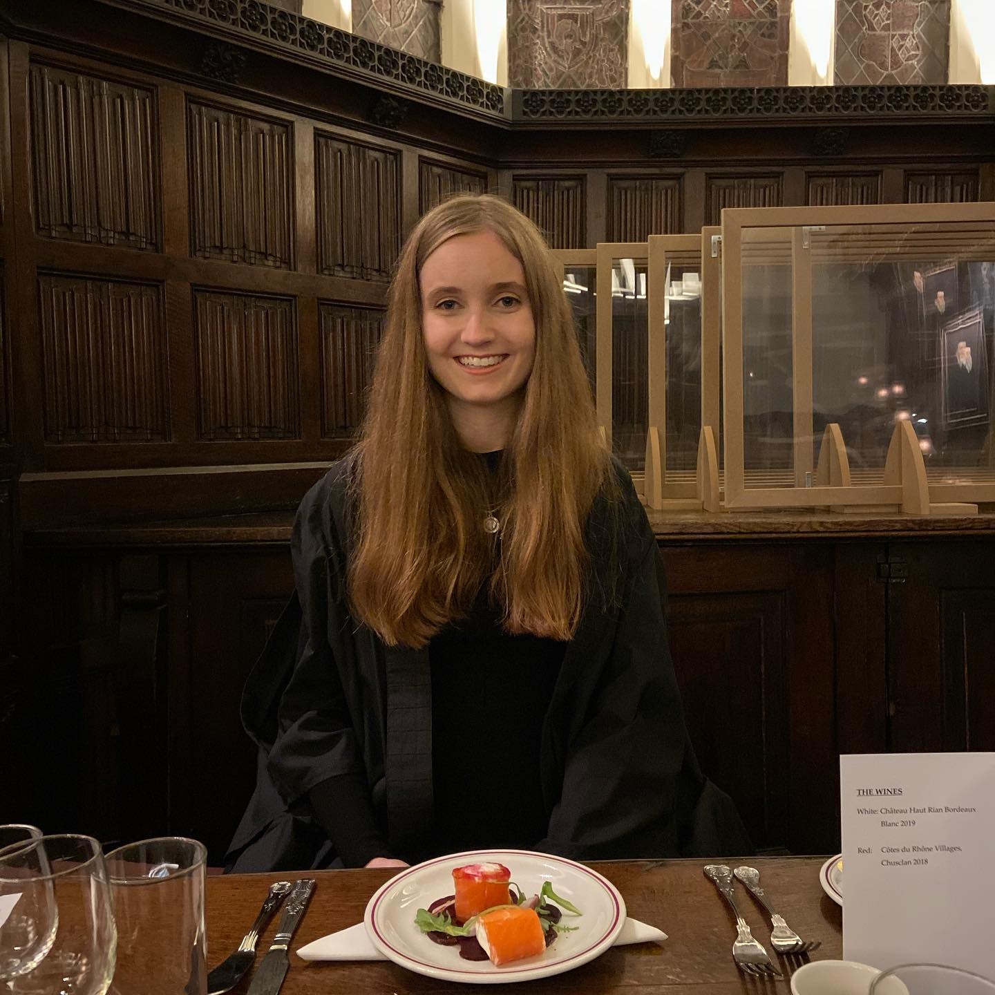 A student in a gown, in front of a plate of food, smiling. She's in front of some old-looking wood-panelled walls