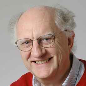 Headshot of John Clarke smiling, wearing glasses, a shirt and a red jumper