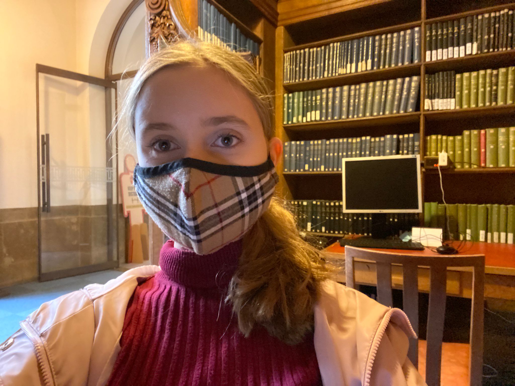 A selfie of Emily, wearing a face mask, with a big bookshelf behind her