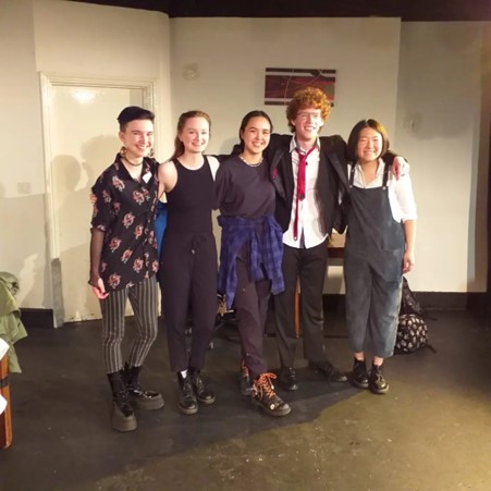 A group of five people on a theatre set, smiling at the camera.