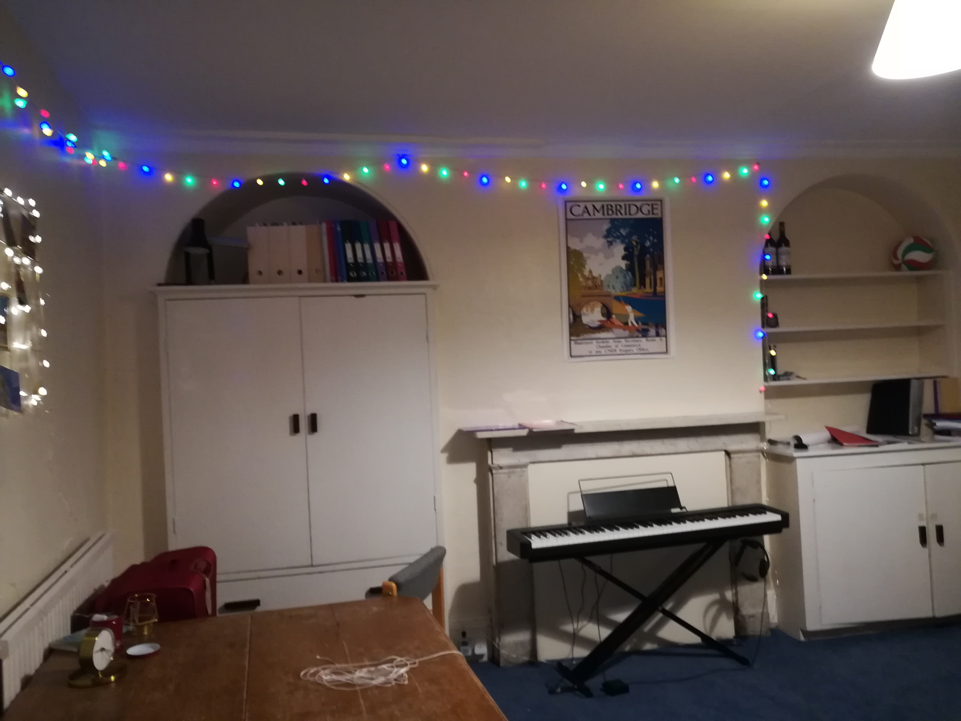 A student room, spacious with fairly lights, a keyboard, a shelf of folders and a poster of Cambridge