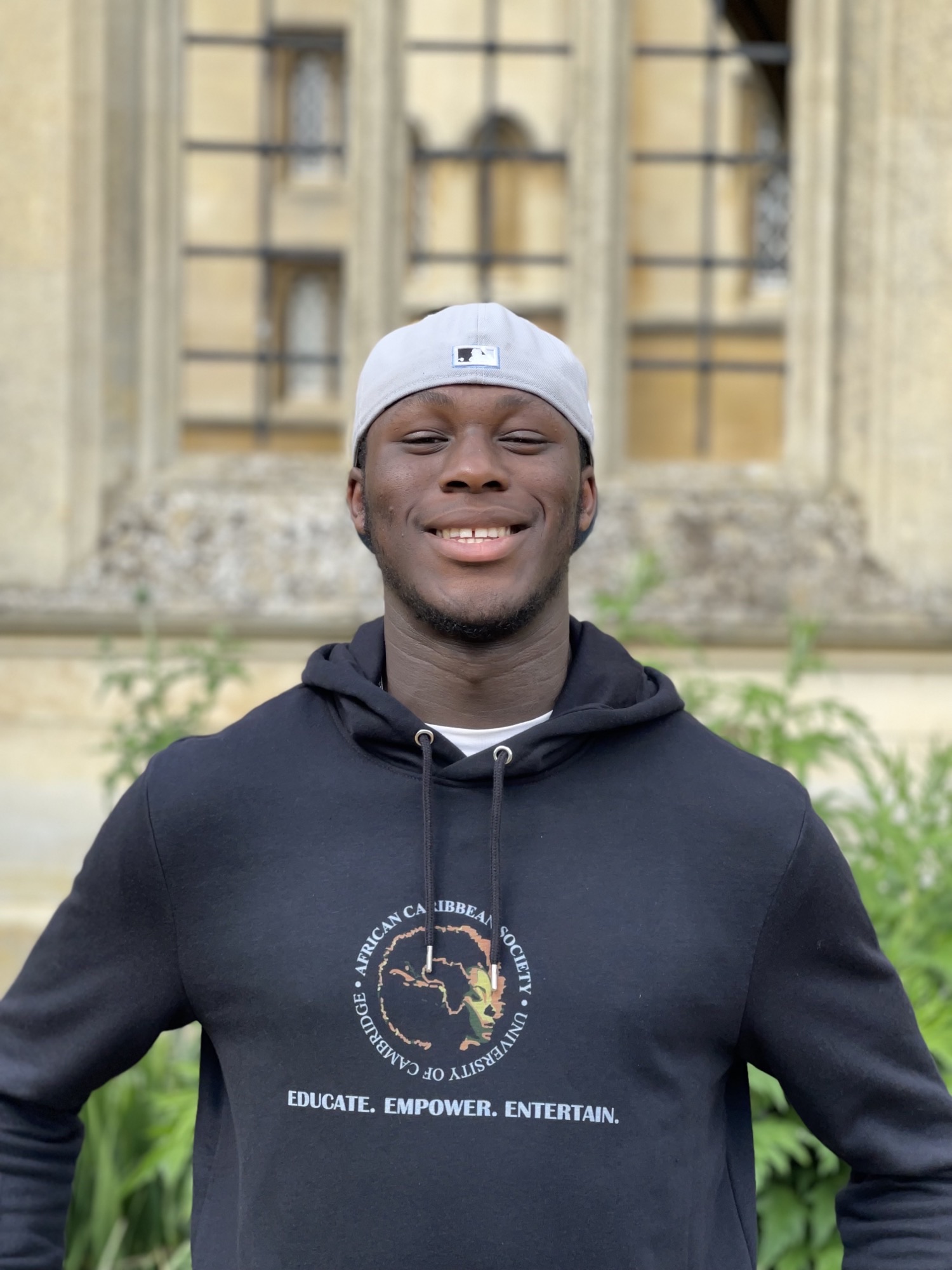 Student wearing a white hat an an afro-caribbean society hoodie, smiles at the camera in front of an old building.