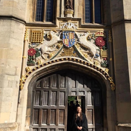 A student stands in front of Christ's main gate, wearing a gown