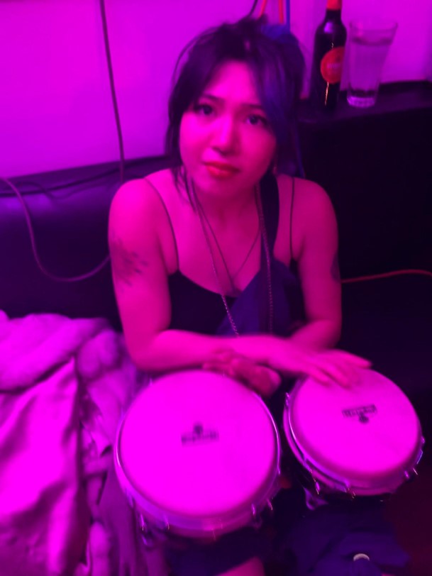 Ari, lit in pink, with some bongos on her lap