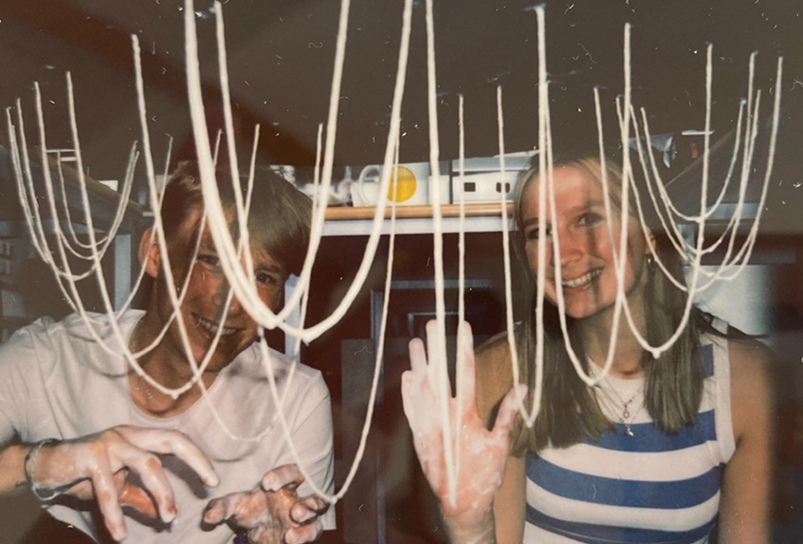 Two students with messy hands, surrounded by strings hanging from a ceiling, wave at the camera.
