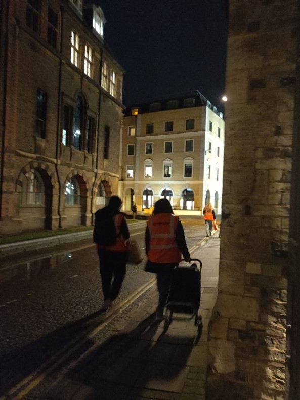 Two students in high-vis jackets, walking down a Cambridge street at night with a trolley of food. 