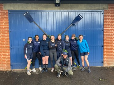 A group of nine people in rowing gear, stood in front of a big blue door with rowing blades on the front