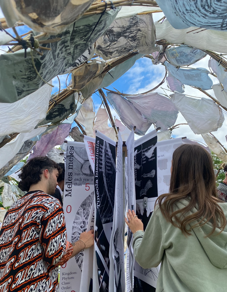 Two students in a structure made from paper/cloth, sticks and cable ties, looking at some sheets of paper