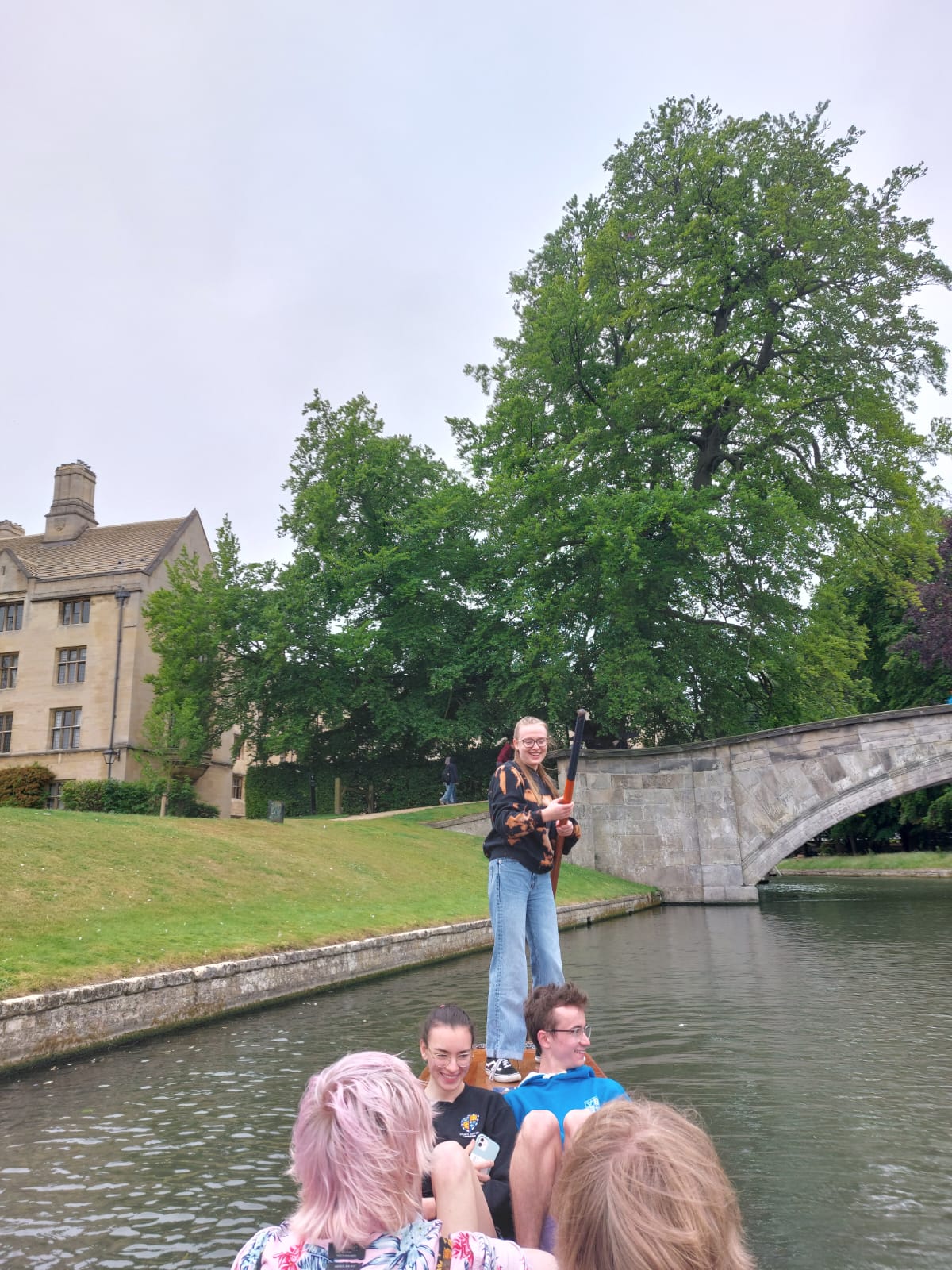 rachel stands on a punt, holding a punting pole, while her friends sit inside and watch the view