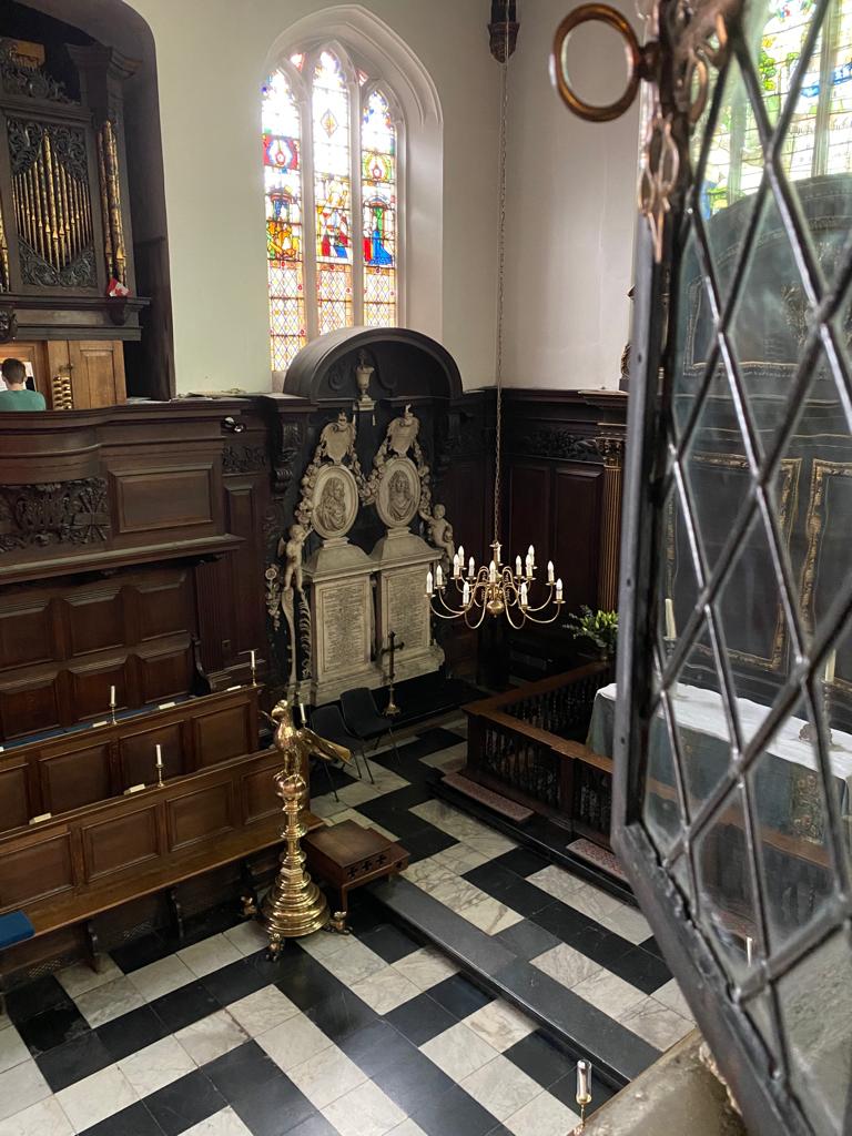 An open window looking out onto Christ's chapel, with a patterned floor, candles hanging from the ceiling, and a monument