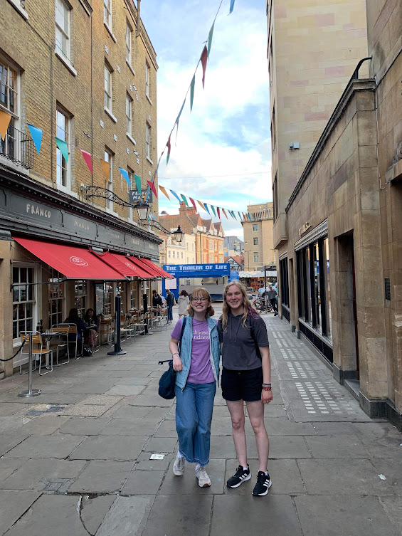 Two students in a street with bunting going accross it, next to a pizza shop