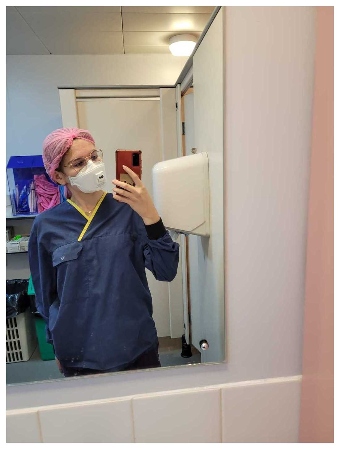 A student in full lab gear, including a face mask and hair net, taking a photo in a mirror