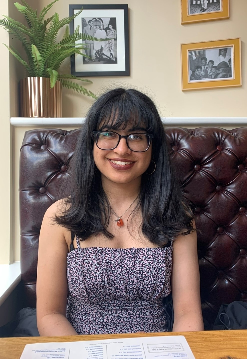 Immi, a young woman with long dark hair and a fringe sat at a table, smiles at the camera