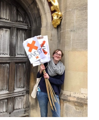 A young woman holding a sign reading 'divest', outside the front gate of Christ's College, Cambridge