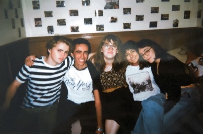 Five students sitting with their arm around each other on a bed in front of a wall covered in polaroids.