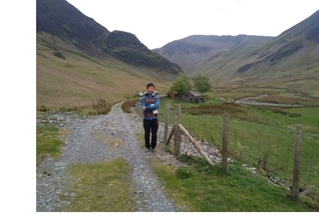 A young man in hiking gear on a small track, with a mountain in the background.