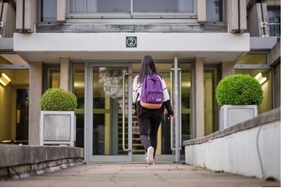 A set of glass double doors in a brutalist concrete building, towards which a woman with a purple backpack is walking (Typewriter building, New Court, Christ's College, Cambridge)