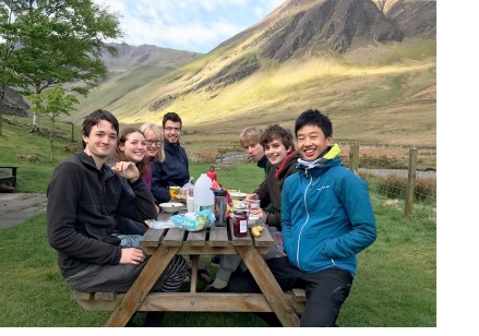 Seven people, in hillwalking clothes and boots, sat around a wooden bench laden with breakfast food, with a mountain in the background.