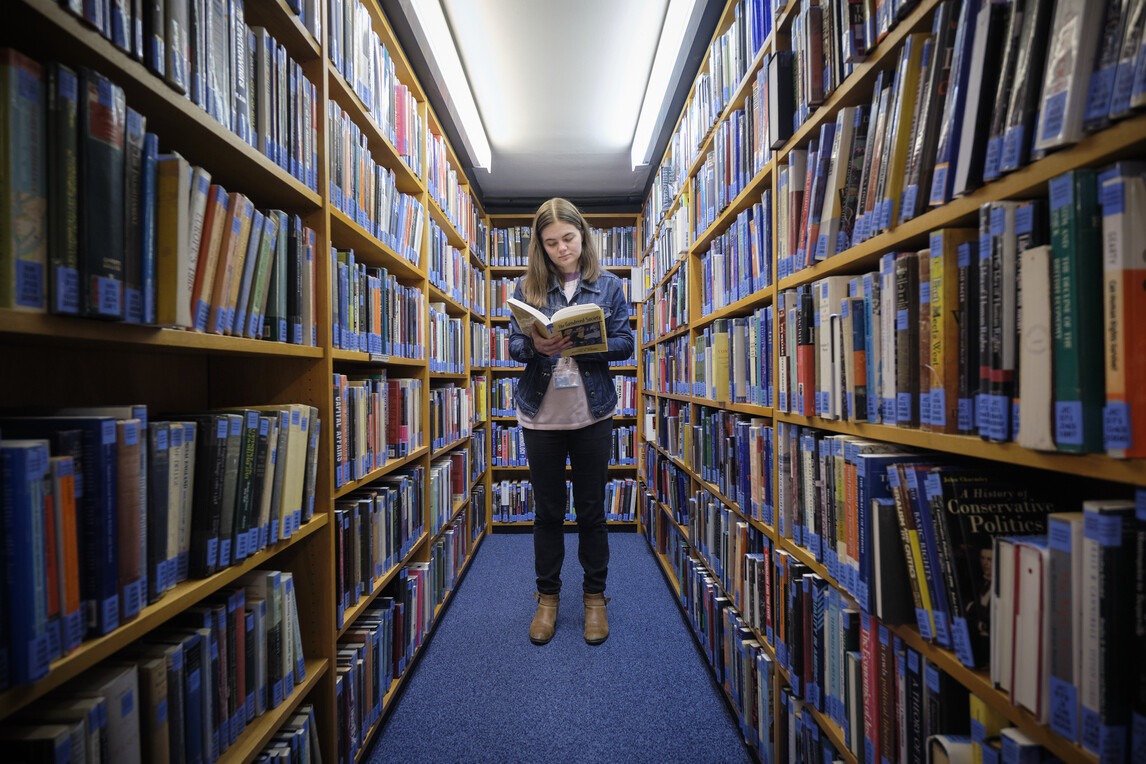 Student reading book between shelves