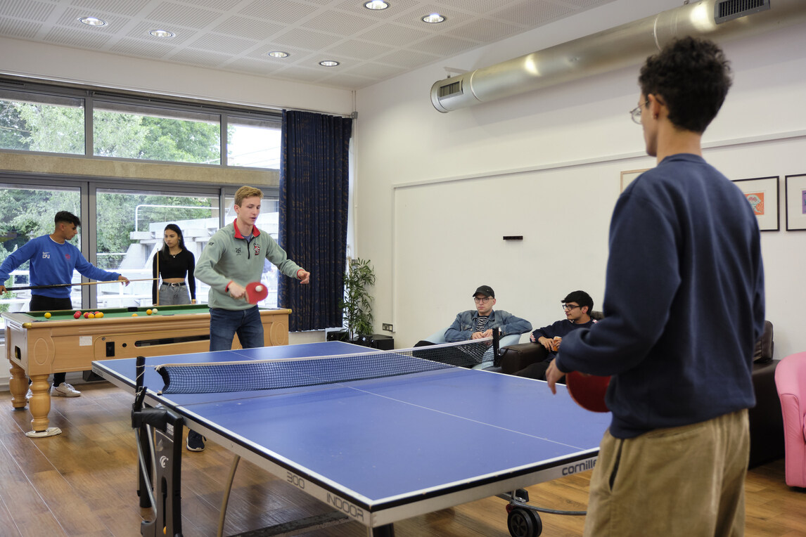 Students playing table tennis with a pool game in background