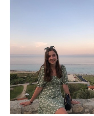 Diana, in a green dress, leaning on a stone wall looking over the sea, at sunset.