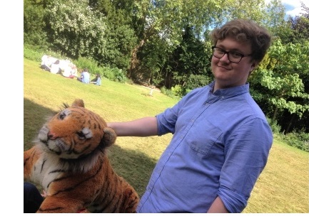 A young man resting his hand on the neck of a toy tiger, in the Fellows' Garden of Christ's College, Cambridge.