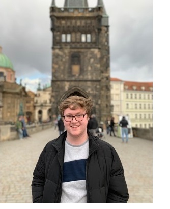 A shot of a young man, smiling at the camera, on Charles Bridge in Prague. The background of the photo is blurred.