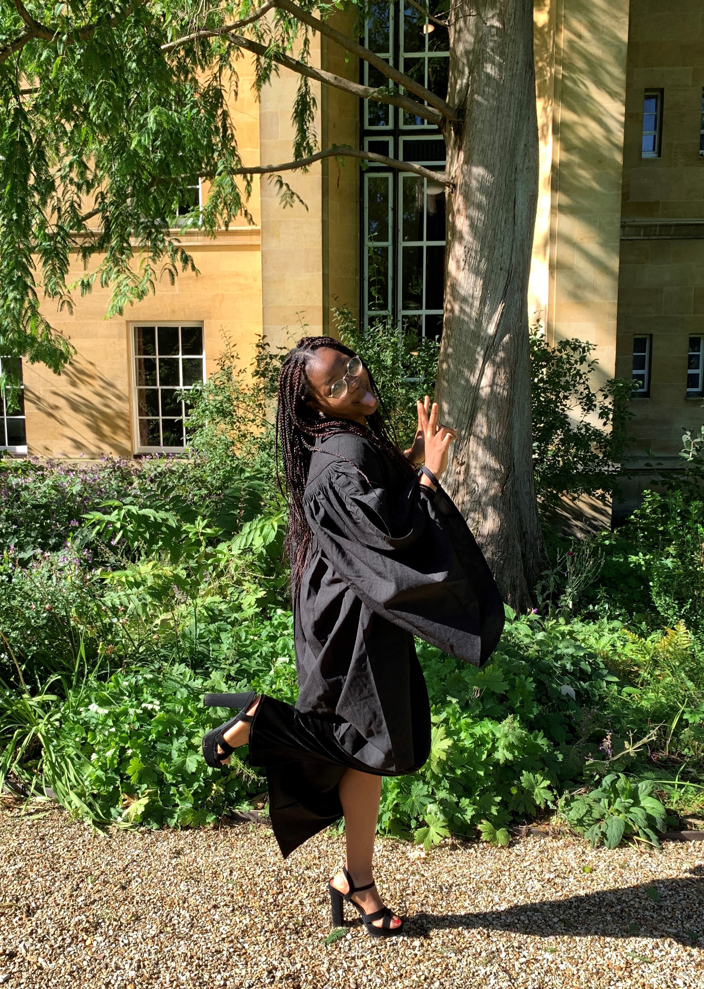 A student in a gown making 'peace signs' and sticking out her tongue, in front of a tree in Third Court