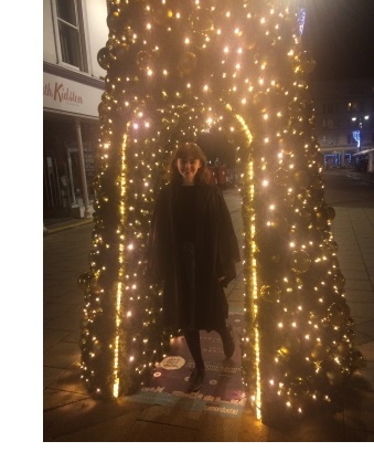 A young woman in formal dress and an academic gown under a large Christmas tree.