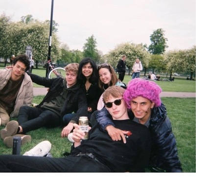 A group of students sprawled out on grass, one of them wearing a pink wig. A man in the centre holds up a can to the camera.
