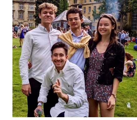 Four students, in smart casual clothes, posing for a photo, with a garden party going on in the background.