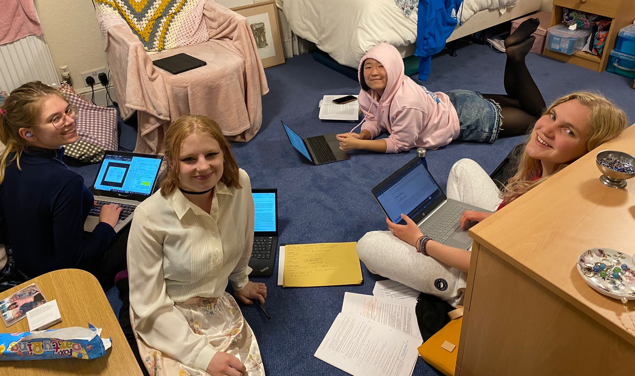 Four students lying on the floor of a slightly chaotic room, with laptops