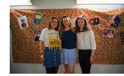 Three young women in casual clothes with their arms around each other, stood in front of a cloth backdrop