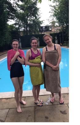 Three young women standing at the side of an outdoor swimming pool, wrapped in towels.