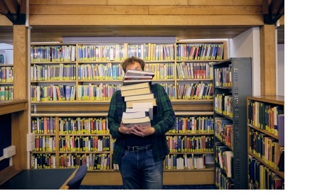 A male student holding a stack of books in front of a library shelf. The top of his right eye - he is wearing round glasses - is just visible above the books.
