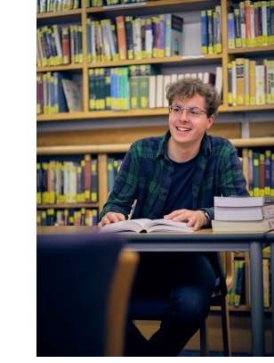 A picture of a male student, in a library, looking up from a book and smiling.