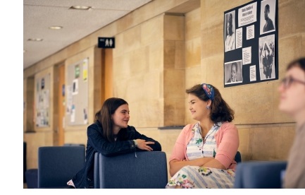 Two female students chatting, sat on sofas in the library corridor of Christ's College, Cambridge.