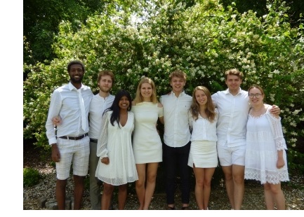 A group of students, dressed in white, posing with their arms around each other in front of a flowering hedge.