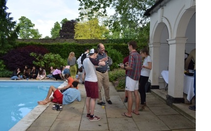A group of students and fellows, with drinks and cake, gathered around the side of the outdoor pool in the Fellows Garden at Christ's College, Cambridge. 