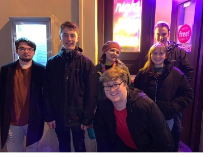 Four young men and two young women posing together under a neon sign.