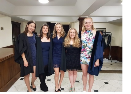 Hannah and a group of friends in blue dresses and academic gowns, attending a formal dinner.