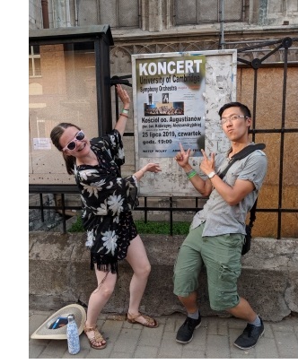 A woman (left) and man (right) pointing at a poster (centre) advertising a concert by the University of Cambridge Symphony Orchestra