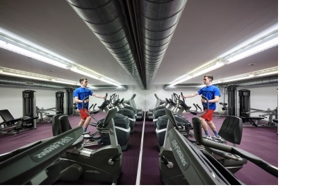 A man, reflected in the mirror spanning the wall, on a treadmill in the gym at Christ's College, Cambridge.