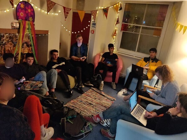 A group of friends in a room with bunting, fairy lights and a rug, using laptops. 