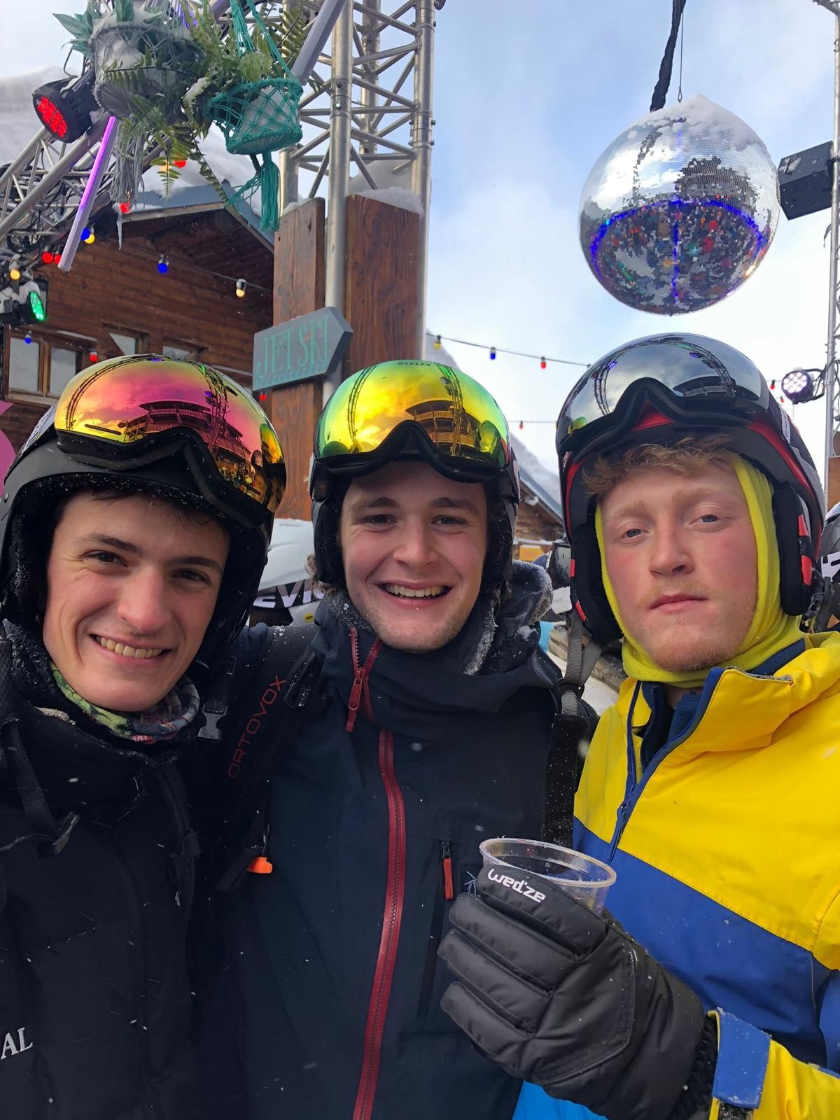 three men in ski gear, one holding a drink, smiling at the camera