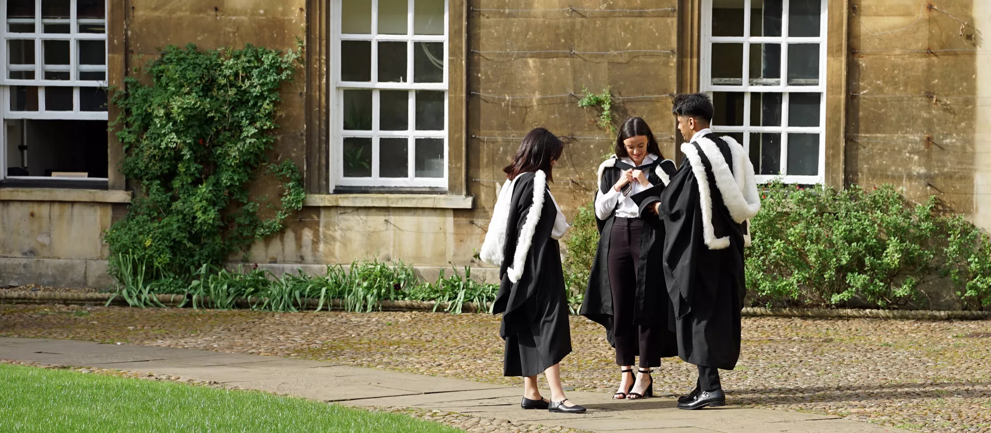 Three robed students stood on grass at their MA graduation ceremony