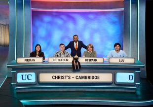 Four people seated in a row in a TV studio, with a fifth person standing behind them