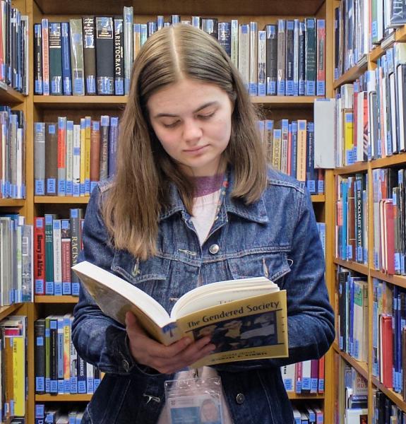Female, standing with open book, reading between bookshelves.
