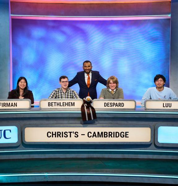 Four people seated in a row in a TV studio, with a fifth person standing behind them